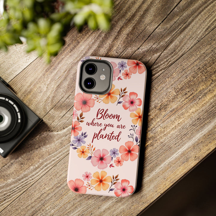 A light pink phone case with a flower garland design that reads Bloom where you are planted surrounded by blooming flowers