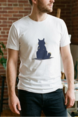 Man wearing a white t-shirt with a graphic design of a cat sitting on a moon, standing in a room with plants and art supplies.