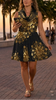 A woman walking on a tropical boardwalk at sunset, wearing a sleeveless black dress with large gold floral mandala art on classic black. Palm trees line the walkway next to the ocean, and other pedestrians are visible in the background.