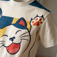 Flat lay of a white t-shirt featuring Laughing Cat Ukiyo-e line art in blue and red accents on a light background, with cat motifs and woodgrain texture for a calm, humorous vibe.