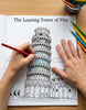 A person's hands are shown coloring in a black and white line art coloring book page of 'The Leaning Tower of Pisa' with a red pencil, on a wooden table with other colored pencils.